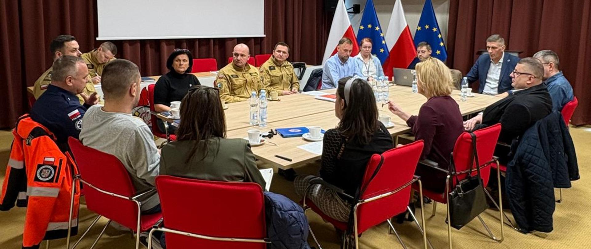 Przy drewnianym stole, na czerwonych fotelach siedzą uczestnicy spotkania. Na sali są beżowa wykładzina i bordowe kotary. Na ścianie po lewej stronie zdjęcia widać biały ekran projektorowy.