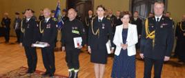 Dzień Strażaka w Rawiczu. Sala widowiskowa w domu kultury. Na tle pododdziału w szeregu stoja strażacy, którzy zostali uhonorowani odznaczeniami. Towarzyszą im pani minister, komendant wojewódzki PSP i komendant powiatowy PSP. 