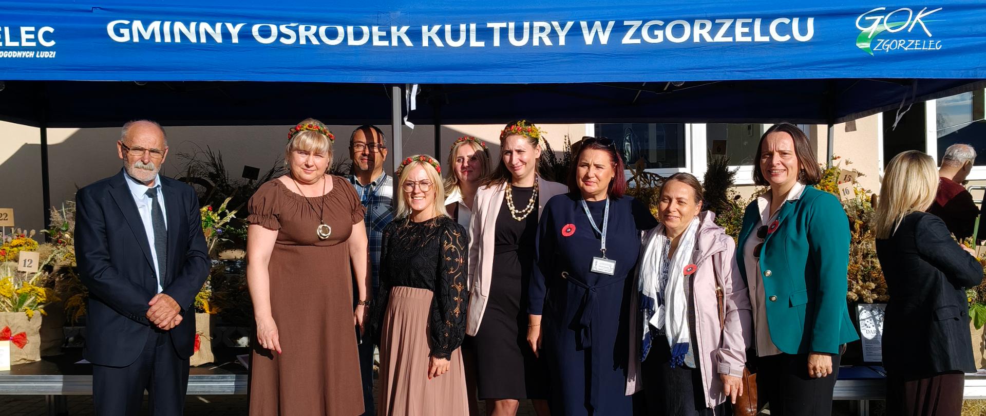 Grupa dziewięciu osób stojących przed stoiskiem wystawienniczym podczas wydarzenia w Zgorzelcu. Osoby pozują do zdjęcia pod niebieskim namiotem z napisem „Gminny Ośrodek Kultury w Zgorzelcu”. W tle widoczne są stoiska z dekoracjami i kwiatami oraz inni uczestnicy wydarzenia.
