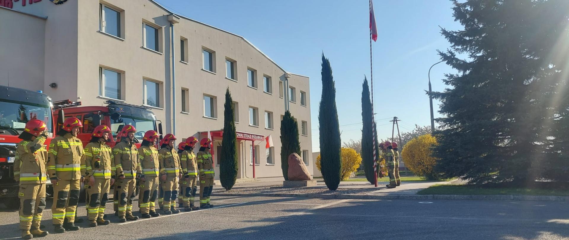 Plac przed budynkiem Komendy Powiatowej Państwowej Straży Pożarnej w Opocznie. Kilkunastu strażaków stojących w dwóch rzędach, wszyscy w ubraniach specjalnych z czerwonymi hełmami. Wszyscy w postawie zasadniczej na baczność. W tle dwa czerwone wozy bojowe strażackie. Trzech strażaków w ubraniach specjalnych w trakcie uroczystego podnoszenia flagi RP na maszt. 