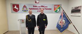 Komendant Miejski Państwowej Straży Pożarnej w Koninie stoi w świetlicy z nowo przyjętym do służby strażakiem. Z boku stoi sztandar komendy. Z tyłu mównica. 