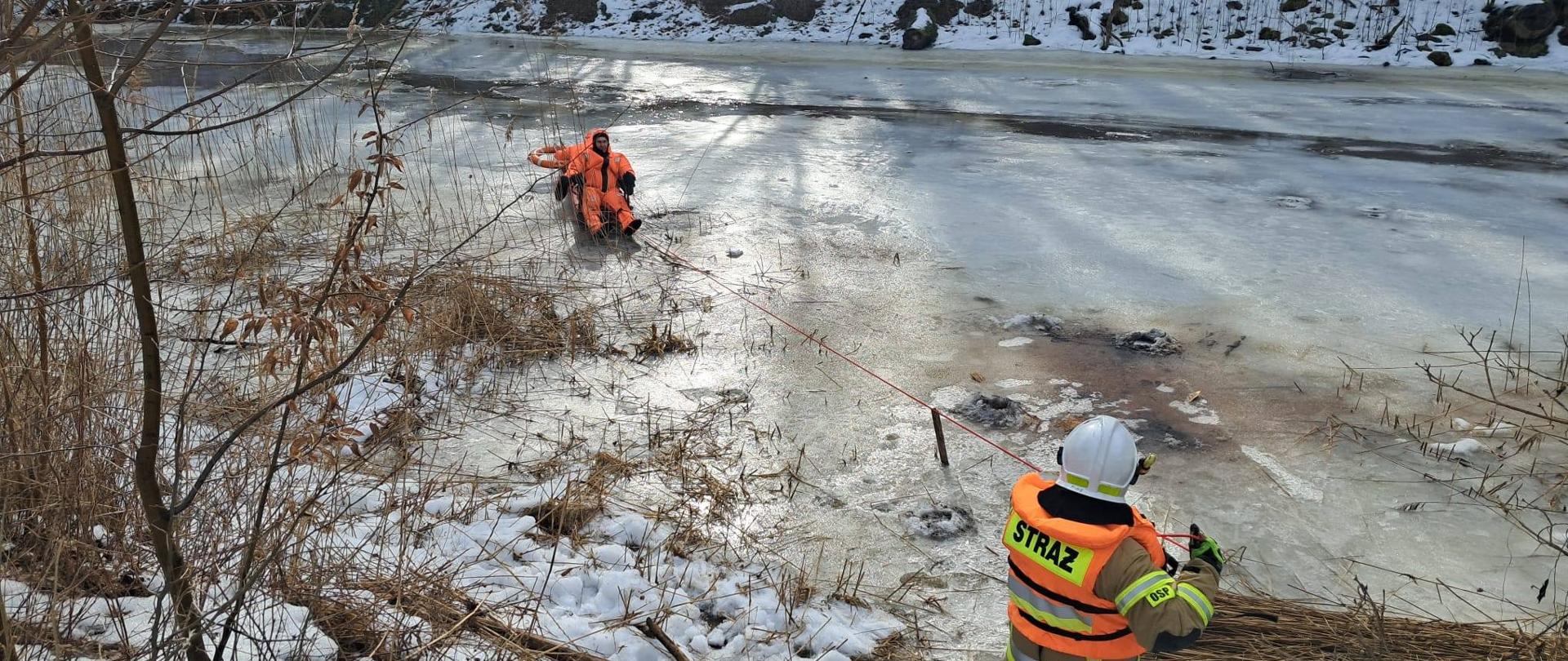 Rzeka skuta lodem. Na rzece w specjalnych kombinezonach dwóch strażaków. Jeden na brzegu trzyma linkę, asekuruje ratowników na lodzie.