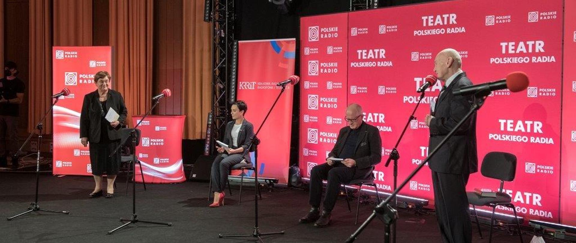 Zdjęcie w poziomie. Na pierwszym planie mężczyzna stoi i przemawia, za nim siedzi kobieta i mężczyzna. Jedna kobieta stoi i patrzy na przemawiającego. W tle banery w kolorze czerwonym