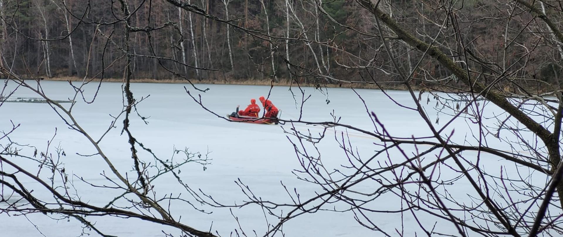Zamarznięta tafla jeziora, w oddali przy wykutym przeręblu dwóch strażaków w skafandrach ochronnych. 