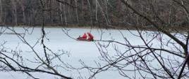 Zamarznięta tafla jeziora, w oddali przy wykutym przeręblu dwóch strażaków w skafandrach ochronnych. 