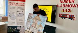 Strażak prezentuje planszę edukacyjną z przekrojem domu, pokazując rozmieszczenie czujników i zagrożeń pożarowych. W tle widoczny jest baner informacyjny „Czujka na straży twojego bezpieczeństwa”, ekran z napisem „Straż Pożarna wita”, walizka z czujnikami oraz napis na ścianie „Europejski Numer Alarmowy 112”. Zdjęcie wykonane w sali edukacyjnej komendy.