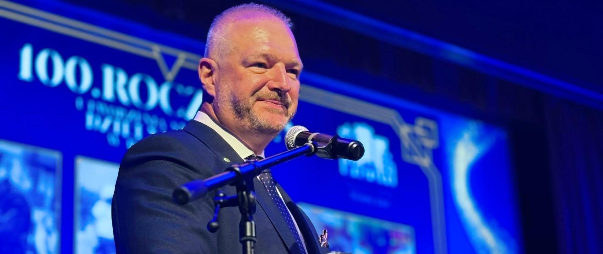 Mężczyzna w garniturze i krawacie stoi przy mównicy, wygłaszając przemówienie. 