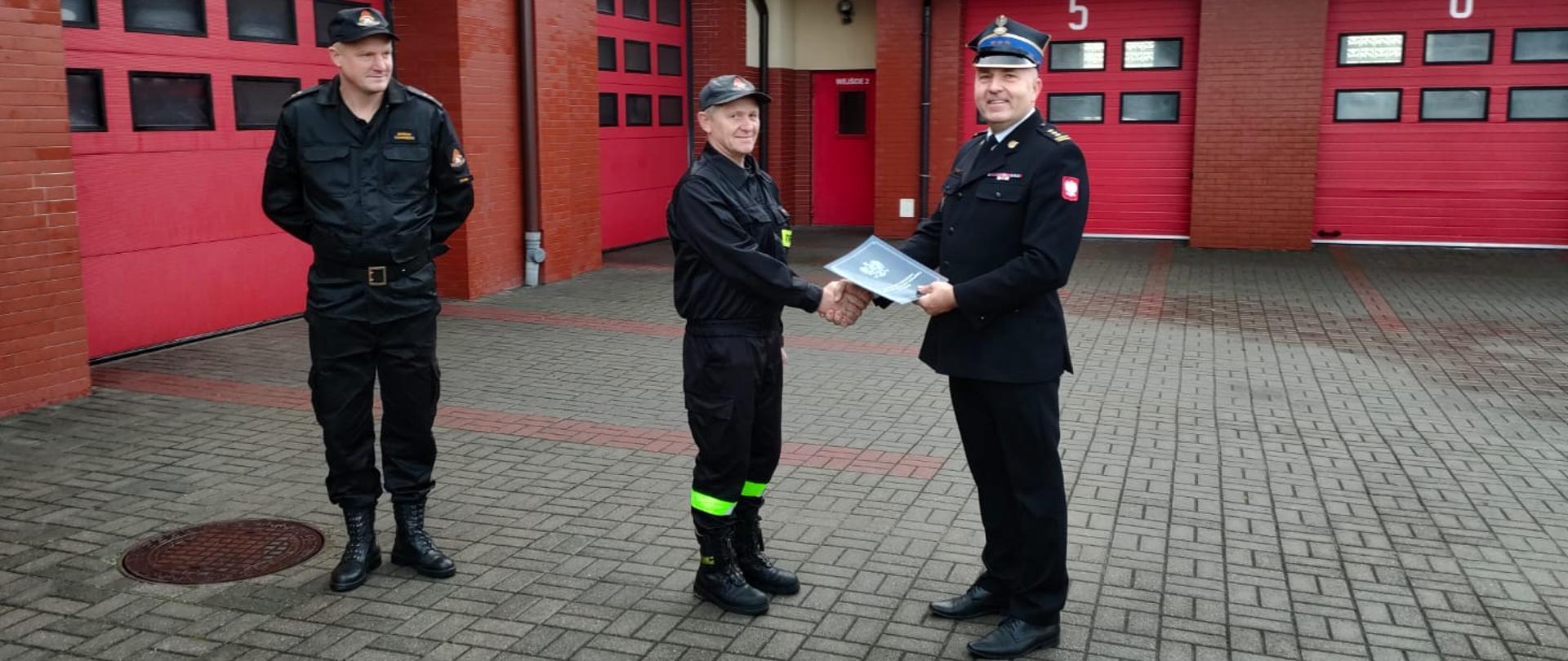 Na zdjęciu na tle garaży JRG Komendant Powiatowy PSP w Rypinie st. bryg. Grzegorz Łydkowski wraz z Zastępcą mł. kpt. Marcinem Czarneckim wręcza list gratulacyjny obchodzącemu jubileusz funkcjonariuszowi asp. sztab. Markowi Murawskiemu
