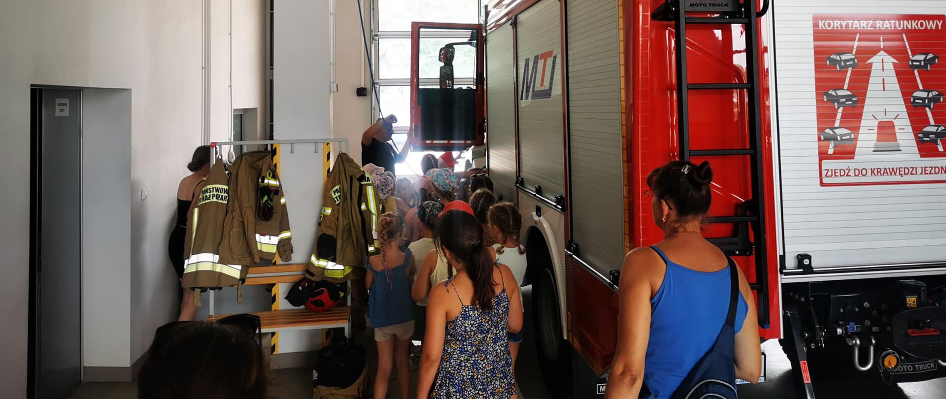Na zdjęciu dzieci przy samochodzie ratowniczo - gaśniczym na wieszaku wiszą ubrania specjalne obok dzieci opiekunowie