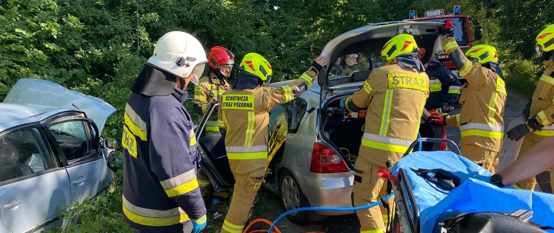 Dwa rozbite samochody widziane z tyłu. Pomiędzy nimi sylwetki strażaków podczas usuwania dachu uszkodzonego pojazdu, jeden strażak trzyma nożyce hydrauliczne i przecina konstrukcję auta.