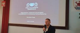 Mężczyzna w garniturze trzyma mikrofon na ekranie jest logo oraz napis Politechnika Gdańska oraz zagrożenia i ochrona od porażeń w budynkach z instalacją fotowoltaiczną obok jest godło Polski oraz Miasta Słupska i zasłony w kolorze flagi Polski. 