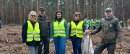 Pracownicy Wojewódzkiego Inspektoratu Ochrony Roślin i Nasiennictwa w Warszawie, Oddziału w Radomiu oraz Regionalnej Dyrekcji Lasów Państwowych w Radomiu pozują do zdjęcia na tle lasu podczas akcji sadzenia drzew. W tle widać więcej uczestników wydarzenia, również w kamizelkach, na leśnym terenie przygotowanym pod nasadzenia.