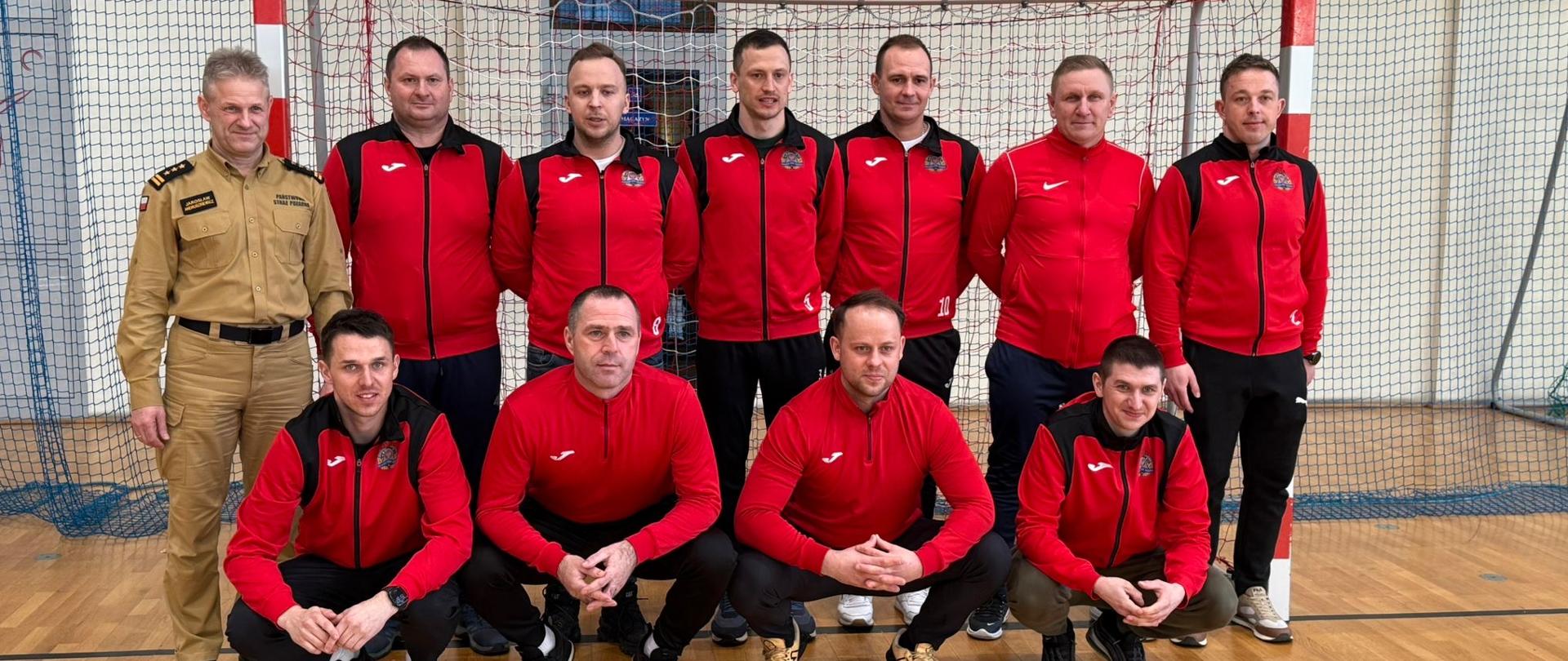 Grupa kilkunastu mężczyzn pozuje do zdjęcia w hali sportowej przed bramką do piłki ręcznej. Większość z nich ma na sobie czerwone bluzy sportowe z czarnymi elementami, a po bokach stoją dwaj mężczyźni w beżowych mundurach. Część osób klęczy z przodu, trzymając dyplomy. Wszyscy ustawieni są w dwóch rzędach na drewnianej podłodze boiska, a w tle widać siatkę bramki i ścianę hali sportowej