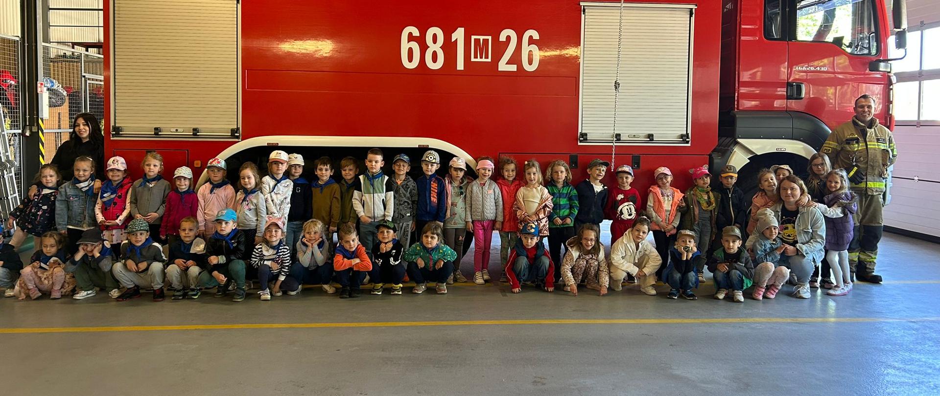 Zdjęcie przedstawia grupę dzieci z przedszkola pozującą do zdjęcia na tle wozu strażackiego. Po prawej stronie zdjęcia stoi strażak w ubraniu specjalnym. Zdjęcie wykonane jest w garażu JRG