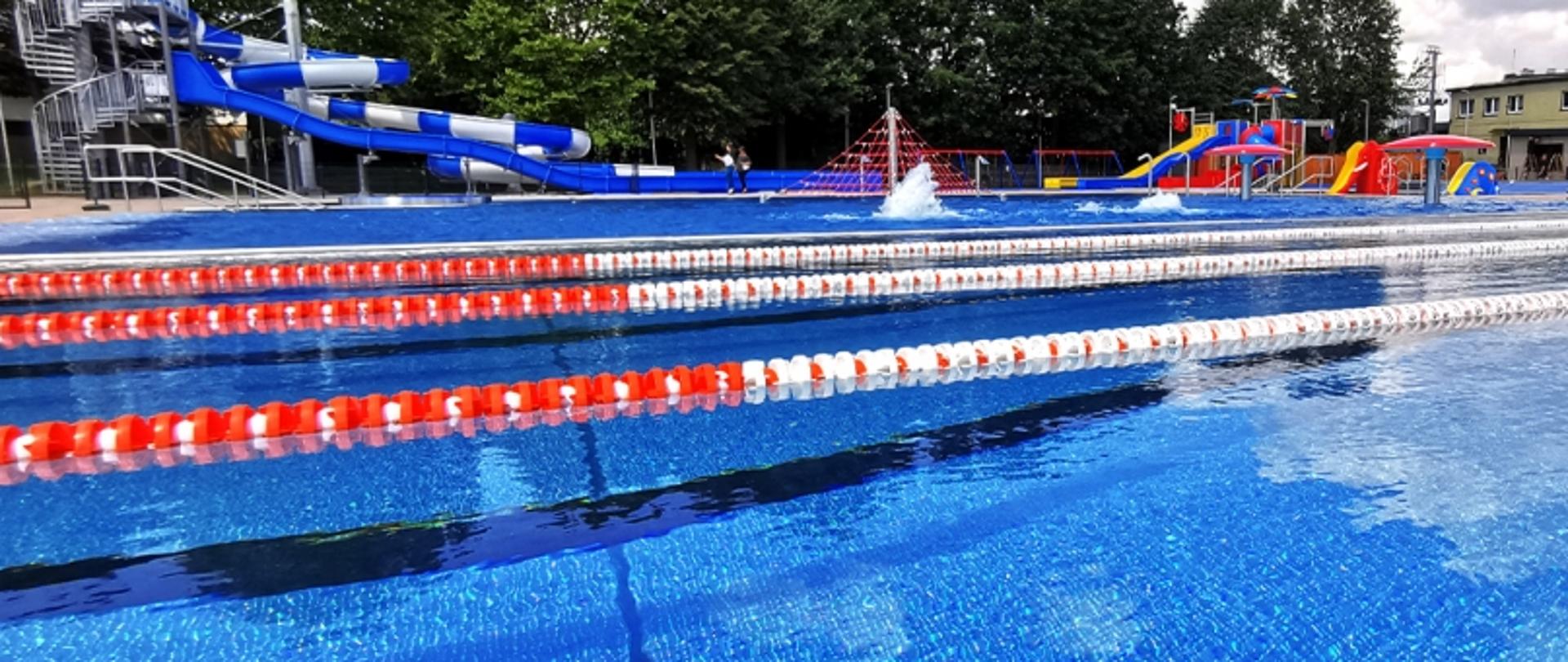 Basen z torami pływackimi oraz wodnym placem zabaw z zjeżdżalniami i siatką wspinaczkową, otoczony drzewami i budynkami.
