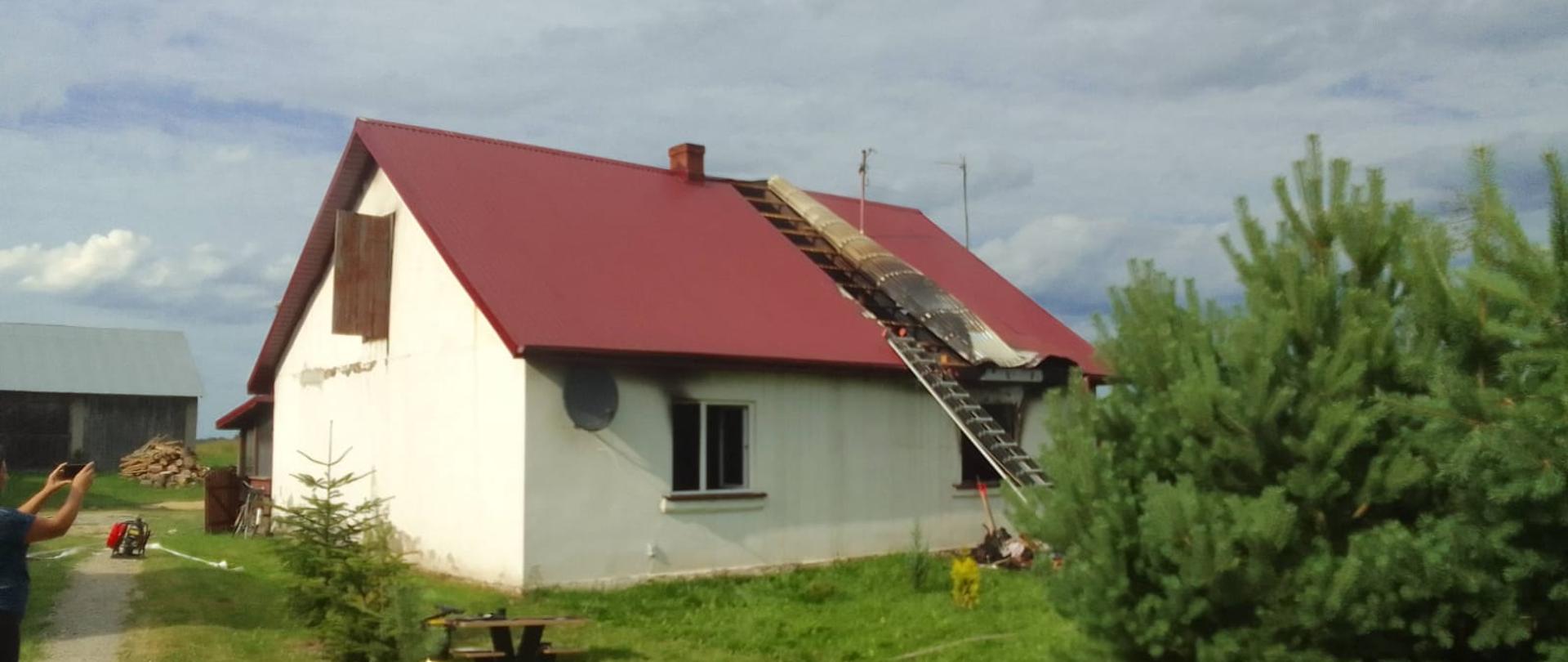 Zdjęcie przedstawia dom jednorodzinny parterowy z czerwonym dachem. Widoczne są ślady pożaru takie jak usunięta blacha z poszycia dachu oraz czarne ślady na otworami okiennymi