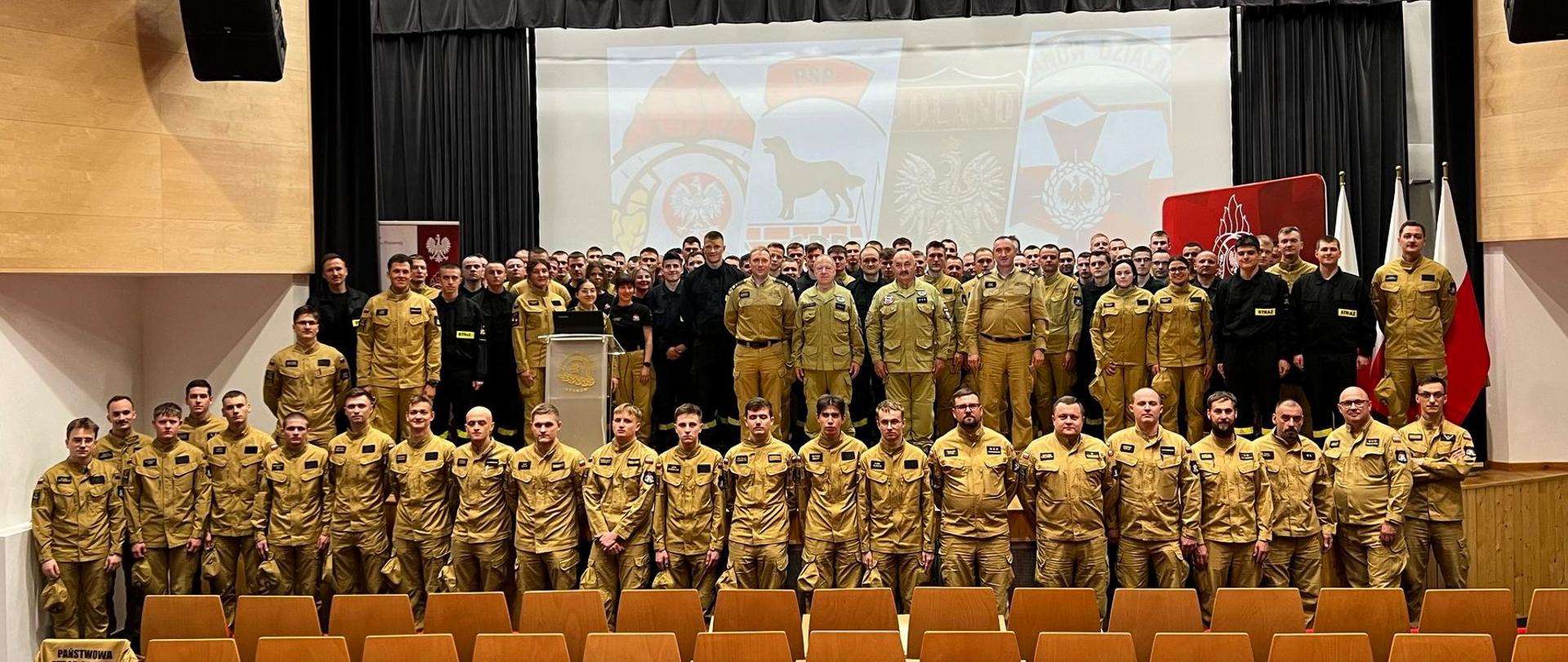 Zdjęcie grupowe wykonane w auli wykładowej Szkoły Aspirantów Państwowej Straży Pożarnej w Krakowie. Na pierwszym planie widocznych jest kilkudziesięciu mężczyzn i kobiet ubranych w beżowe mundury strażackie — to słuchacze i funkcjonariusze szkoły. Stoją w kilku rzędach, zajmując całą szerokość sceny i przestrzeni przed nią. W tle, na ekranie, wyświetlone są emblematy związane z PSP, w tym godło Polski, wizerunek psa ratowniczego oraz różne oznaczenia grup ratowniczych. Po prawej stronie widoczne są trzy stojące flagi: biało-czerwona flaga Polski oraz dwie flagi służbowe. Na środku sceny znajduje się mównica z herbem PSP, a wokół niej zgromadzeni są prelegenci i przedstawiciele kadry, również w mundurach. Uczestnicy wydarzenia stoją z powagą, niektórzy uśmiechają się do kamery. Aula jest dobrze oświetlona i wypełniona rzędem pustych krzeseł na pierwszym planie. Atmosfera zdjęcia jest uroczysta i profesjonalna — odzwierciedla ważne wydarzenie edukacyjne w środowisku służb ratowniczych.