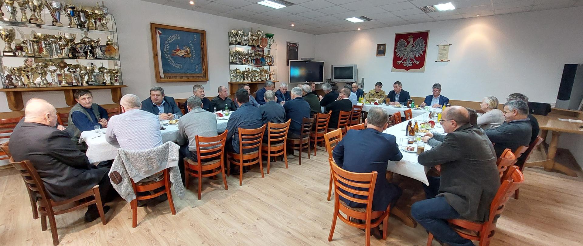 Kolorowa fotografia wykonana w pomieszczeniu, przedstawia zebranie kilkudziesięciu cywilnych osób siedzących przy długich stołach. Jedna osoba za stołem siedzi w jasnym mundurze służbowym strażaka Państwowej Straży Pożarnej. Wokół jasne ściany pomieszczenia, na których wiszą gabloty z pucharami, obrazy, Godło Polski i dyplomy. 