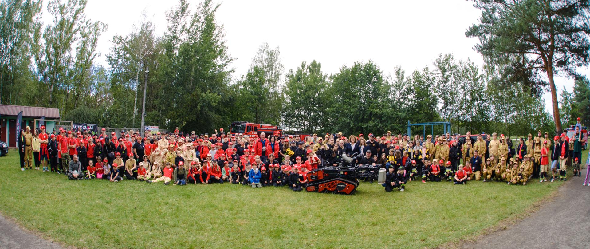 Bardzo duże, panoramiczne zdjęcie grupowe (wykonane obiektywem szerokokątnym) setek uczestników, w większości dzieci i młodzieży w jednolitych czerwonych strojach i czapeczkach, siedzących i stojących na dużej łące otoczonej lasem. Pośrodku grupy stoi nowoczesny, gąsienicowy robot gaśniczy. W tle widoczne są czerwone samochody strażackie.