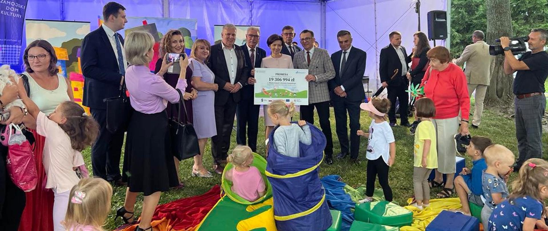 Minister trzyma czek. Obok inni uczestnicy spotkania. Widać dzieci.