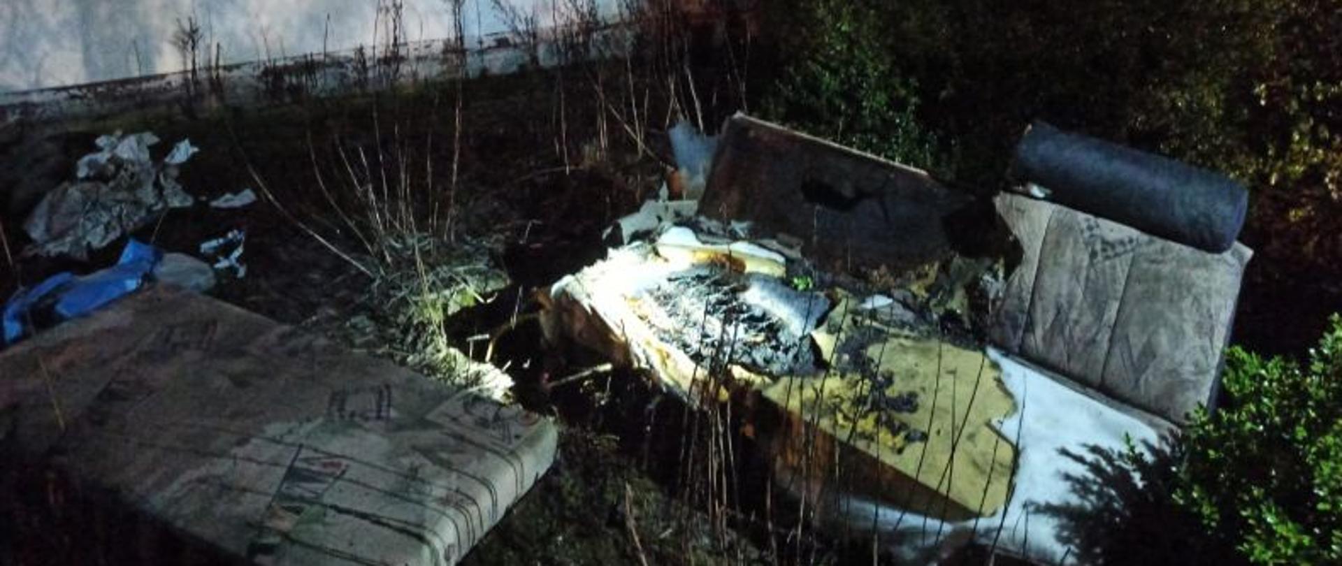 Zdjęcie wykonane w nocy i oświetlone latarkami i najaśnicą strażacką. Przedstawia spalone elementy wnętrza pomieszczenia objętego pożarem. Elementy leżą przy budynku. Budynek jest koloru białego. Tło stanowią drzewka iglaste.