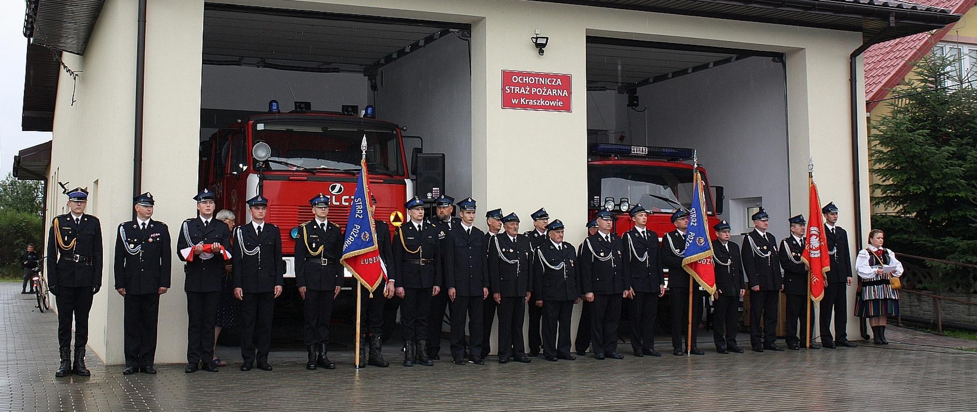 poczty sztandarowe stojące w szeregu przed budynkiem remizy, w budynku otwarte są drzwi garażowe a wewnątrz stoją samochody pożarnicze