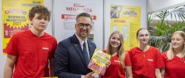 Minister odwiedza stoisko promocyjne. Wspólnie z przedstawicielami ekspozycji pozuje do zdjęcia przy stole, na którym znajdują się materiały informacyjne, modele zwierząt, przypinki oraz gadżety edukacyjne. W tle widoczne są balony, fotografie i banery wystawiennicze.