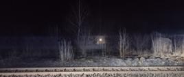 Zdjęcie w porze nocnej przy torach kolejowych. Oświetlenie sztuczne. Widać teren wraz ze spaloną trawą.