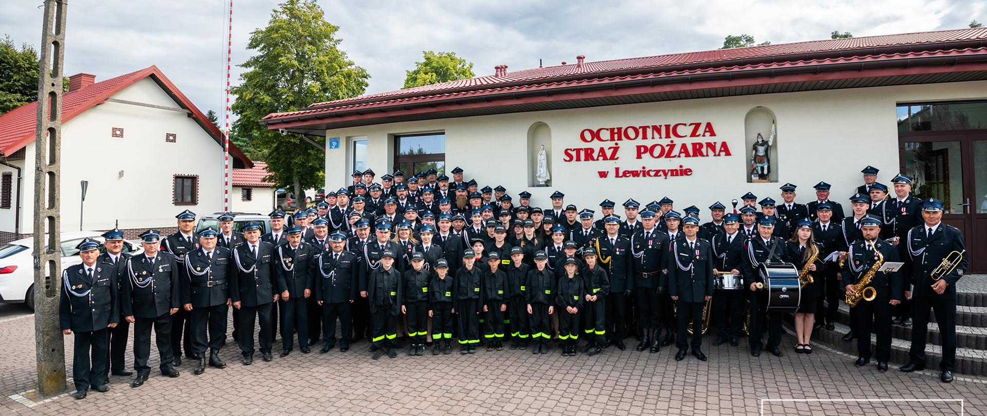 Na zdjęciu grupa dzieci z MDP oraz strażaków ochotników i orkiestry strażackiej na tle budynku remizy