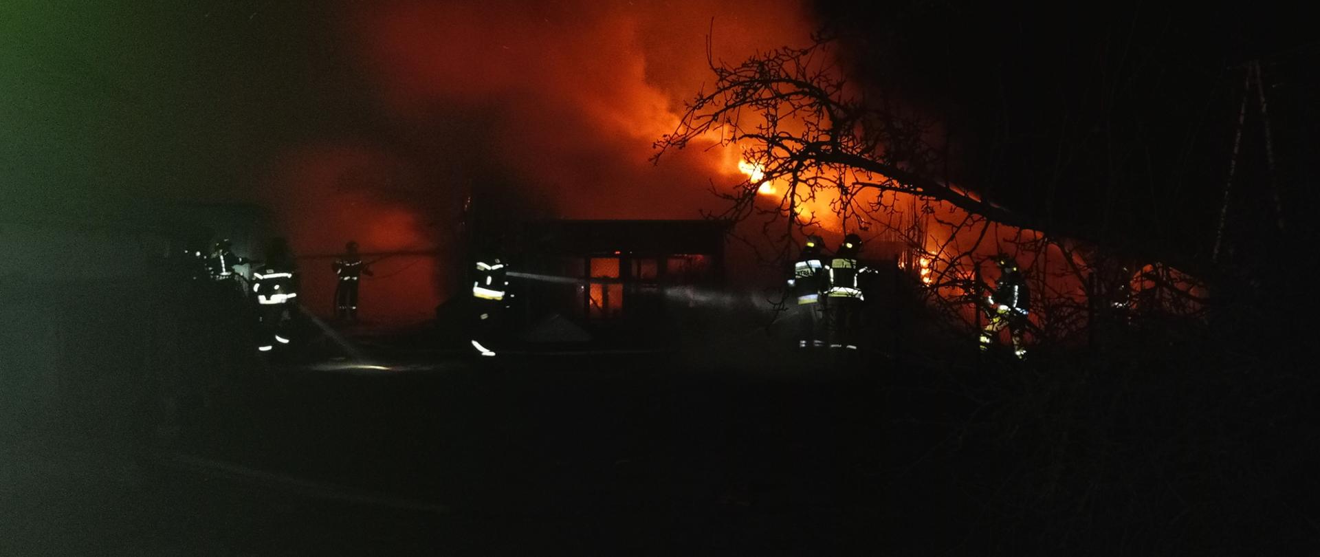 Strażacy wokół palącego się budynku, widać płomienie. Zdjęcie wykonane w porze nocnej 