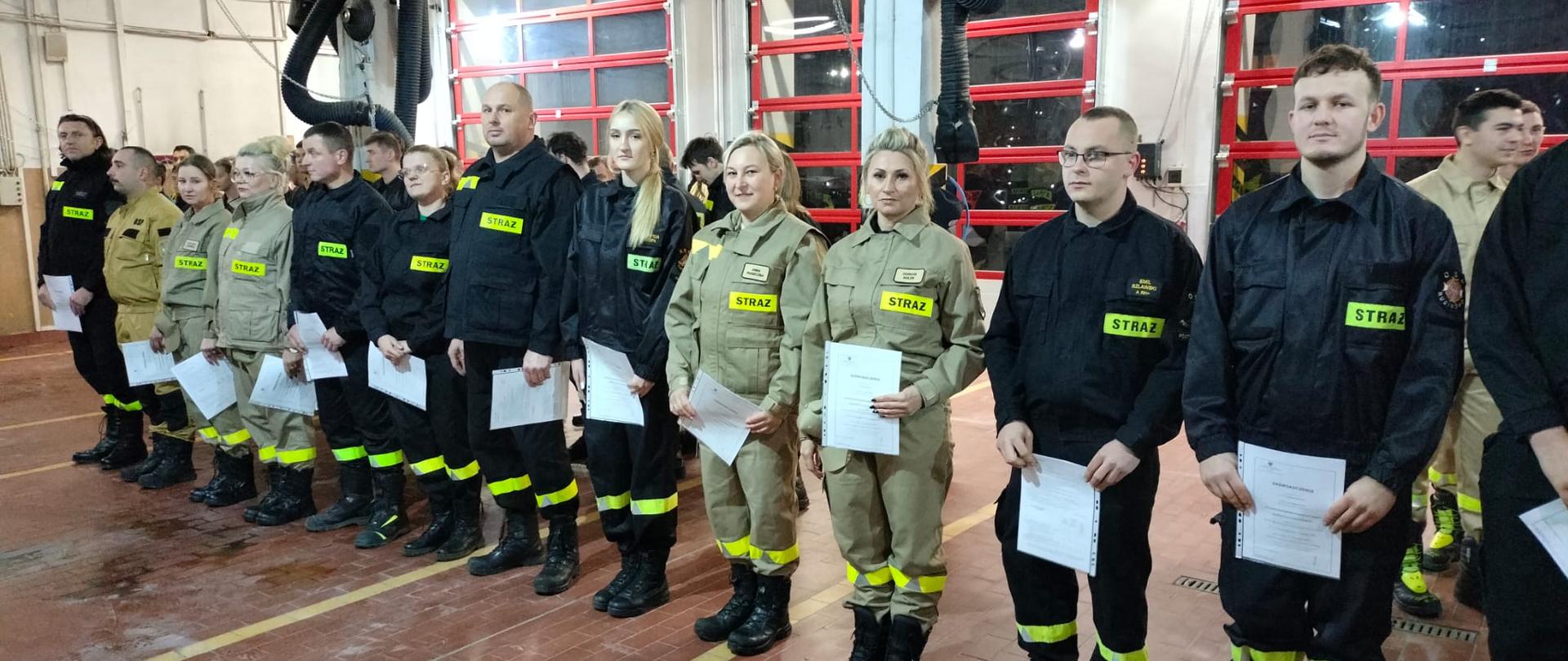 Grupa strażaków w umundurowaniu ochronnym stoi w szeregu w remizie, trzymając dokumenty, podczas uroczystej zbiórki.