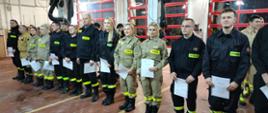 Grupa strażaków w umundurowaniu ochronnym stoi w szeregu w remizie, trzymając dokumenty, podczas uroczystej zbiórki.