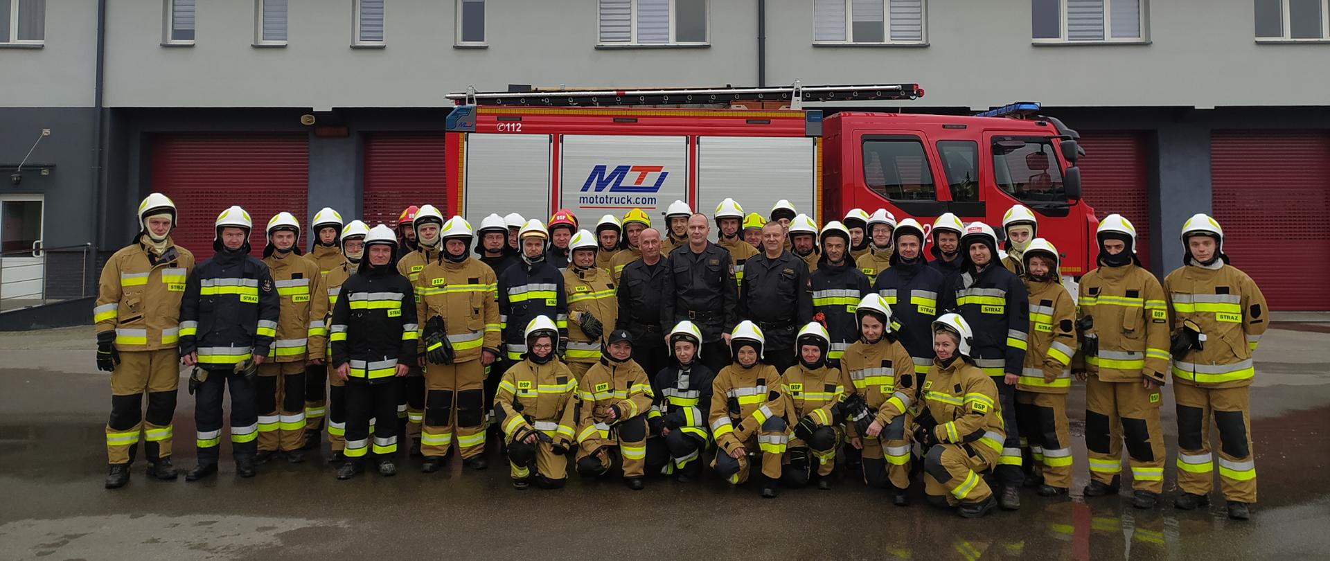 Zdjęcie przedstawia grupę 35 strażaków, którzy ukończyli kurs podstawowy dla OSP oraz w środku trzech funkcjonariuszy - oficerów PSP. Strażacy ochotnicy ubrani są w mundury bojowe, w hełmach białych, natomiast mundury w kolorze piaskowy i część w kolorze czarnym. Strażacy ustawieni w dwuszeregu, w półkolu na tle garaży Komendy Powiatowej PSP oraz na tle czerwonego samochodu ratowniczo – gaśniczego