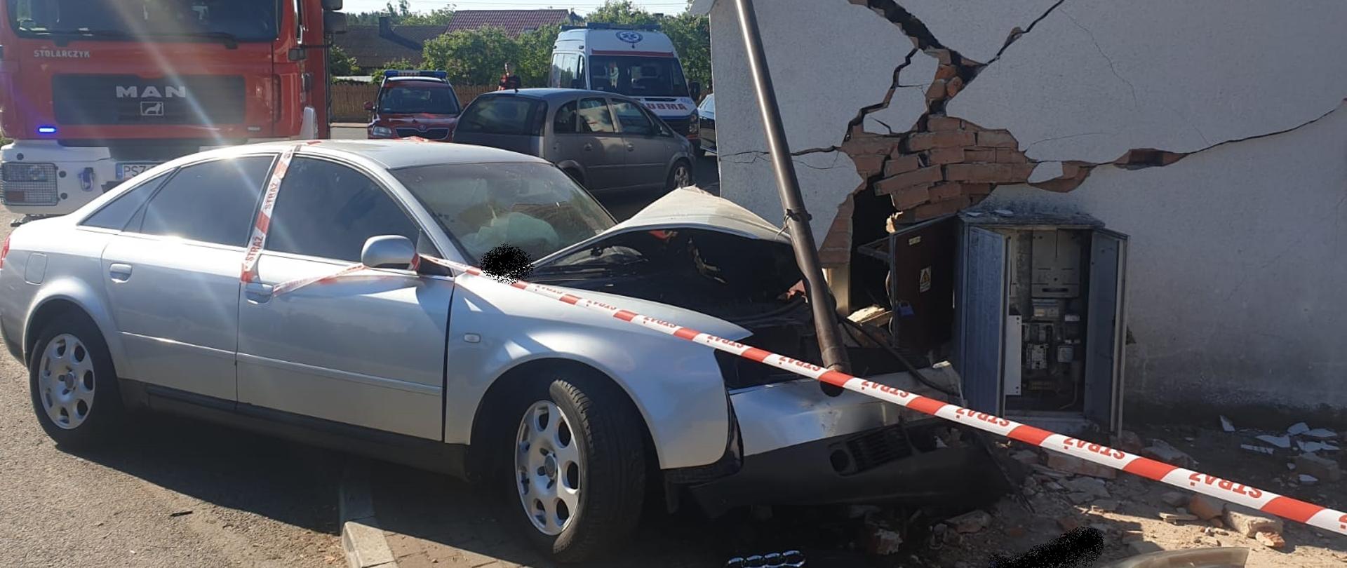 Widać uszkodzony przód samochodu od przodu, uszkodzona ściana budynku, w tle samochód straży