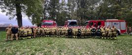 grupa strażaków stojących na trawie a w tle pojazdy pożarnicze
