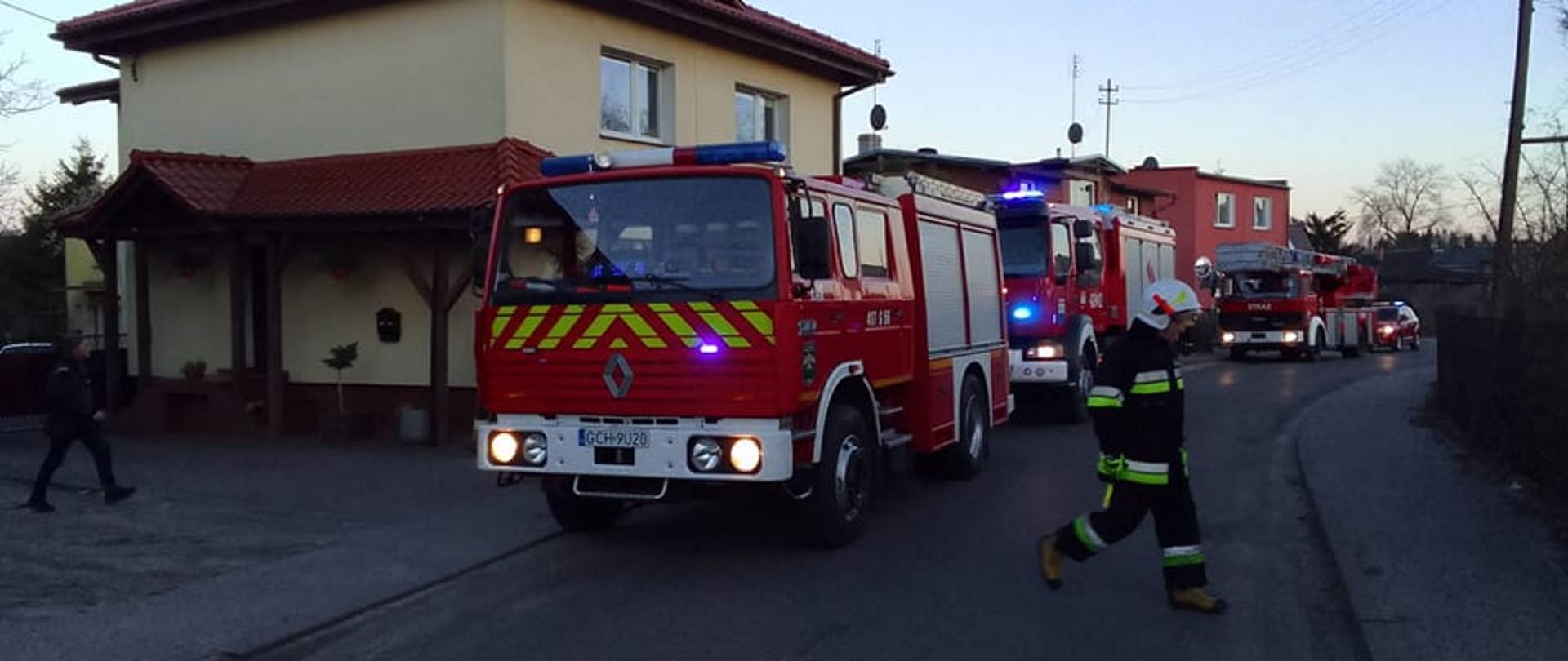 Na ulicy przed budynkiem mieszkalnym zaparkowane są 4 wozy strażackie w włączonymi sygnałami świetlnymi. Przed wozami biegnie strażak uczestniczący w zdarzeniu.