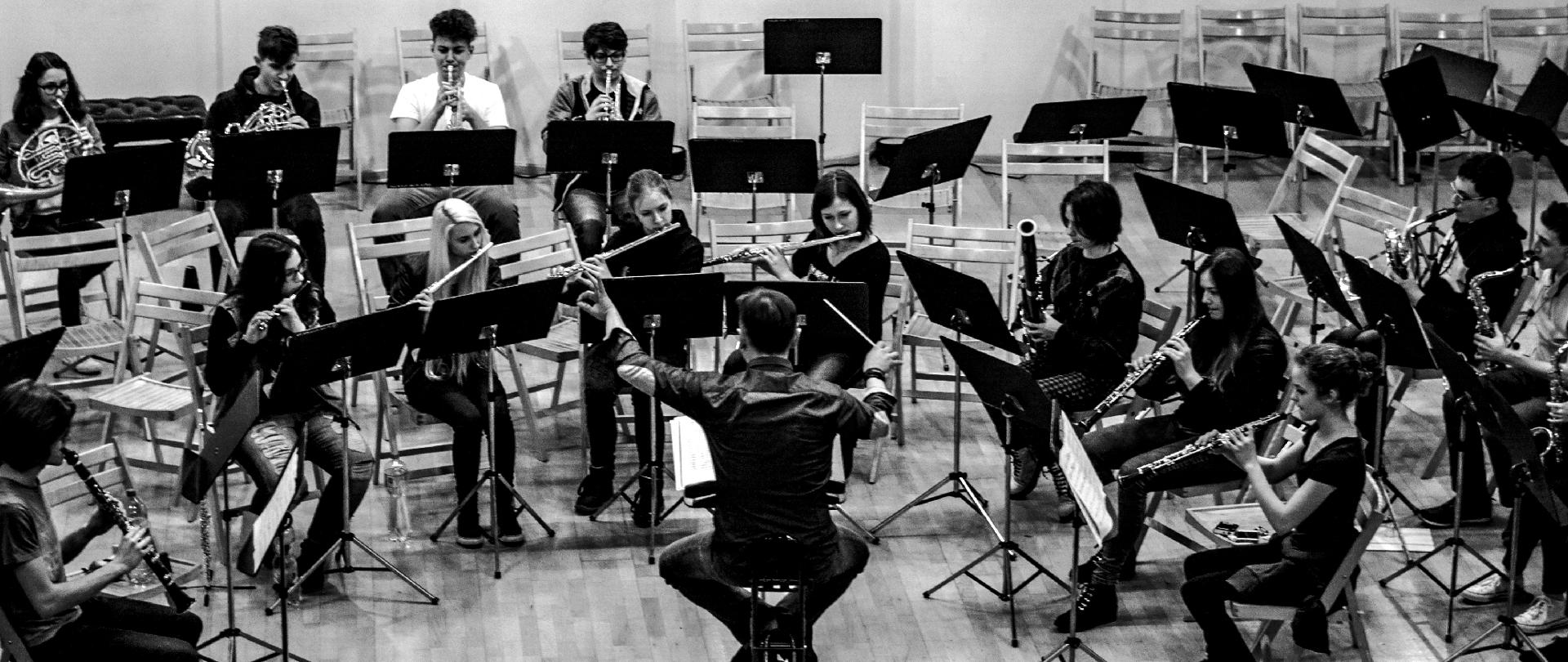 Czarno-białe zdjęcie przedstawiające koncert orkiestry dętej w sali koncertowej ZPSM - uczniowie grający na instrumentach dętych oraz dyrygent