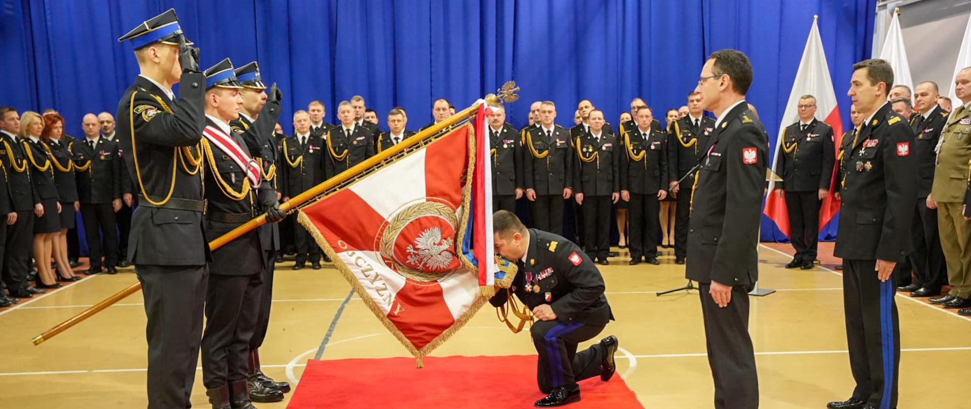 Na sali gimnastycznej stoją funkcjonariusze i osoby cywilne podczas uroczystej zbiórki, na środku sali poczet sztandarowy 3 strażaków PSP ze sztandarem, dotychczasowy komendant wojewódzki PSP klękając całuje sztandar, po jego lewej stronie stoi przyjmujący obowiązki śląskiego komendanta wojewódzkiego, a za nimi komendant główny PSP