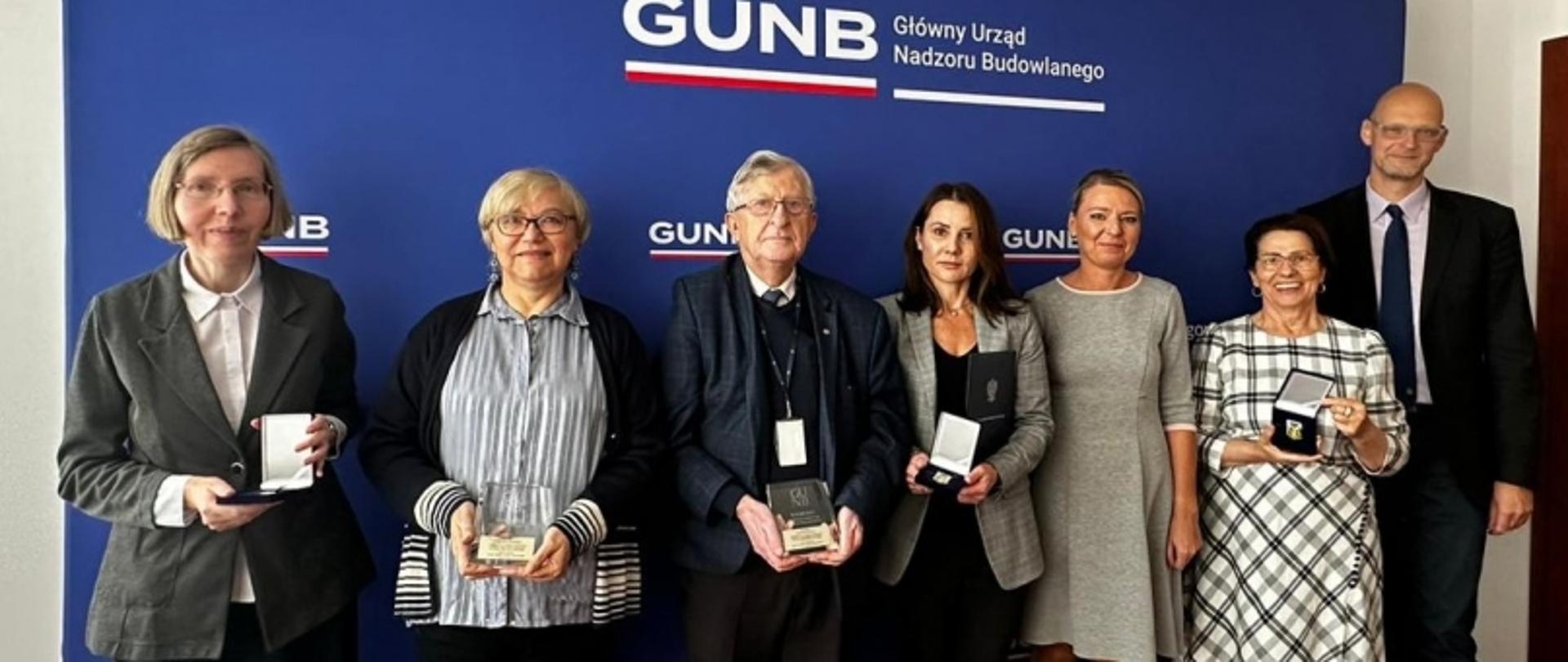 Group of seven people standing in front of a blue backdrop with the text "GUNB Główny Urząd Nadzoru Budowlanego," each holding an award or medal.