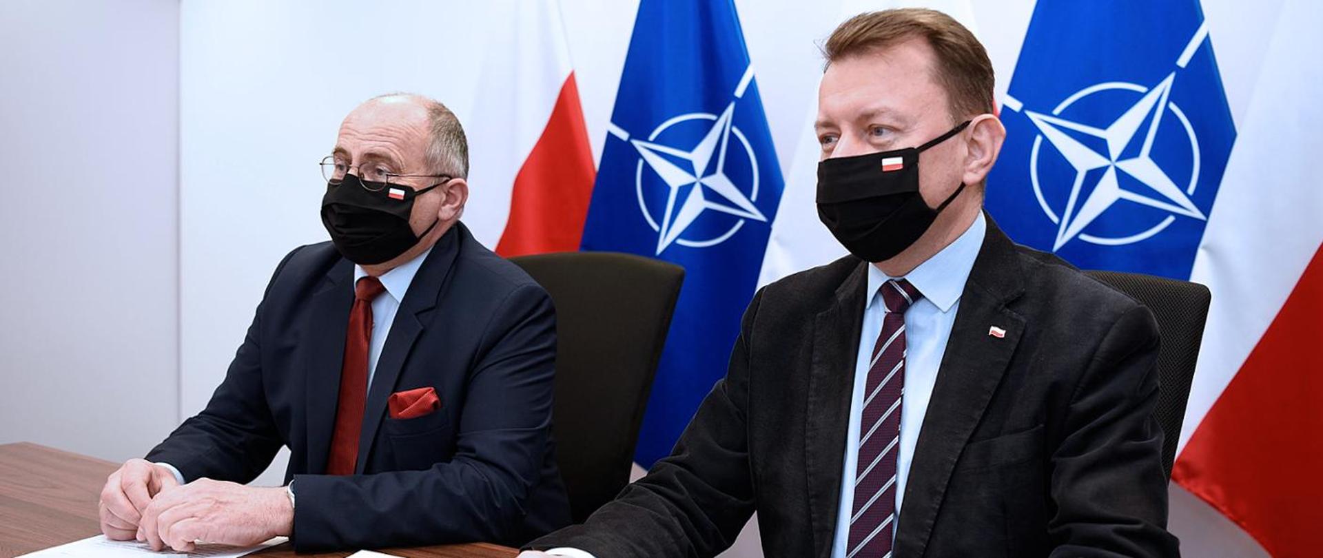 Ministrowie Rau i Błaszczak siedzą za stołem na tle flag Polski i NATO