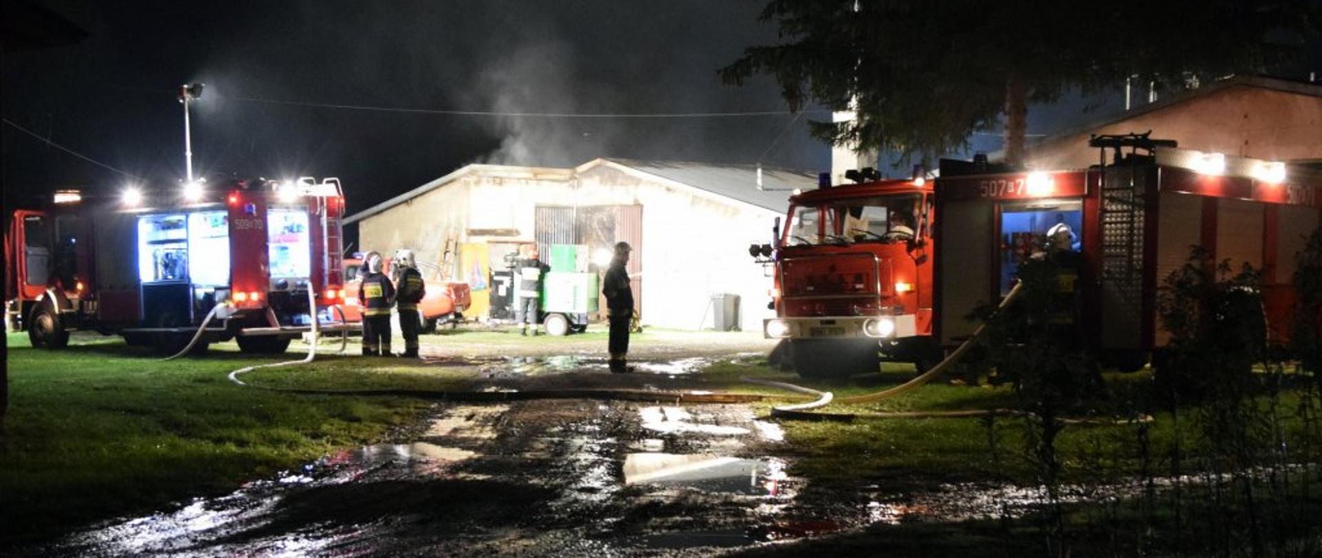 Zdjęcie zrobione w nocy. Na zdjęciu miejsce prowadzenia akcji gaśniczej. Po lewej i prawej stronie stoją oświetlone samochody strażackie. w centrum wydać dymiącą się fermę drobiu. 
