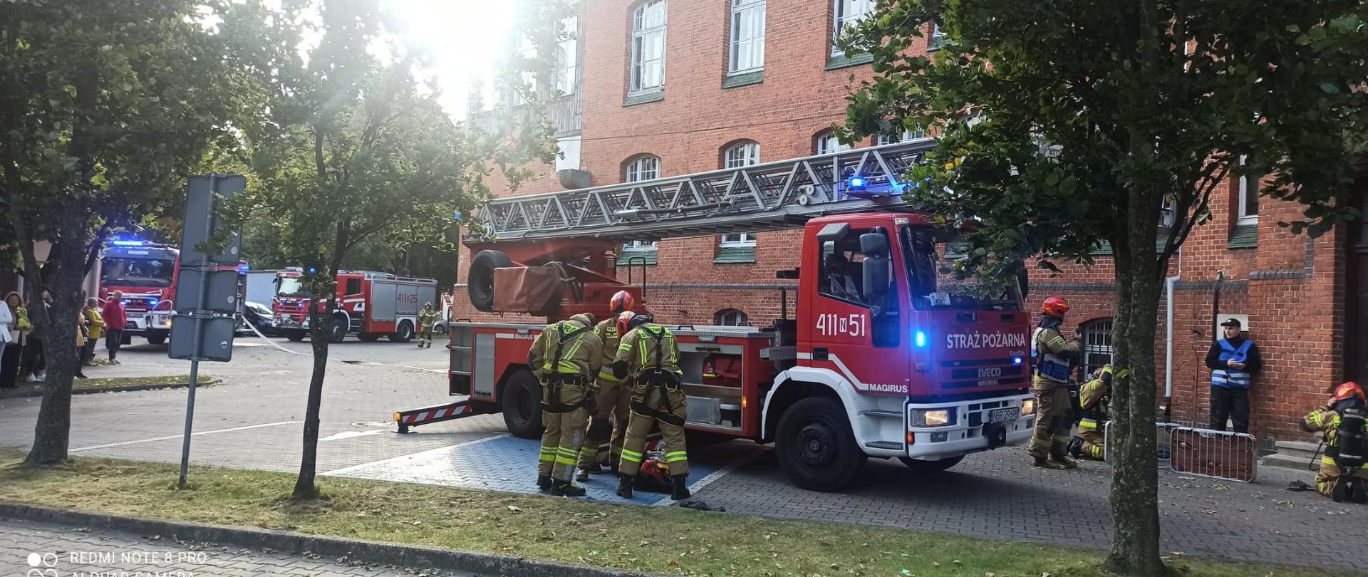 Na zdjęciu samochód drabina strażacka, przy samochodzie strażacy. Na tle budynku z czerwonej cegły. Z lewej w oddali dwa samochody strażackie.