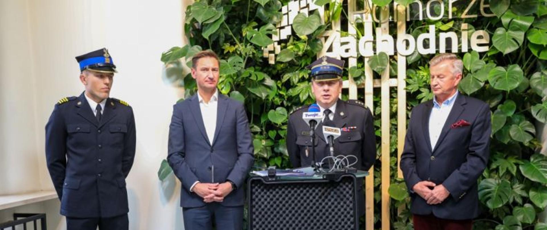 Zdjęcie przedstawia mówiącego do mikrofonu Komendanta Powiatowego PSP Białogard. Po jego bokach stoją Marszałek oraz Członek Zarządu Województwa Zachodniopomorskiego. Po lewej stronie zdjęcia stoi strażak również w galowym ubraniu