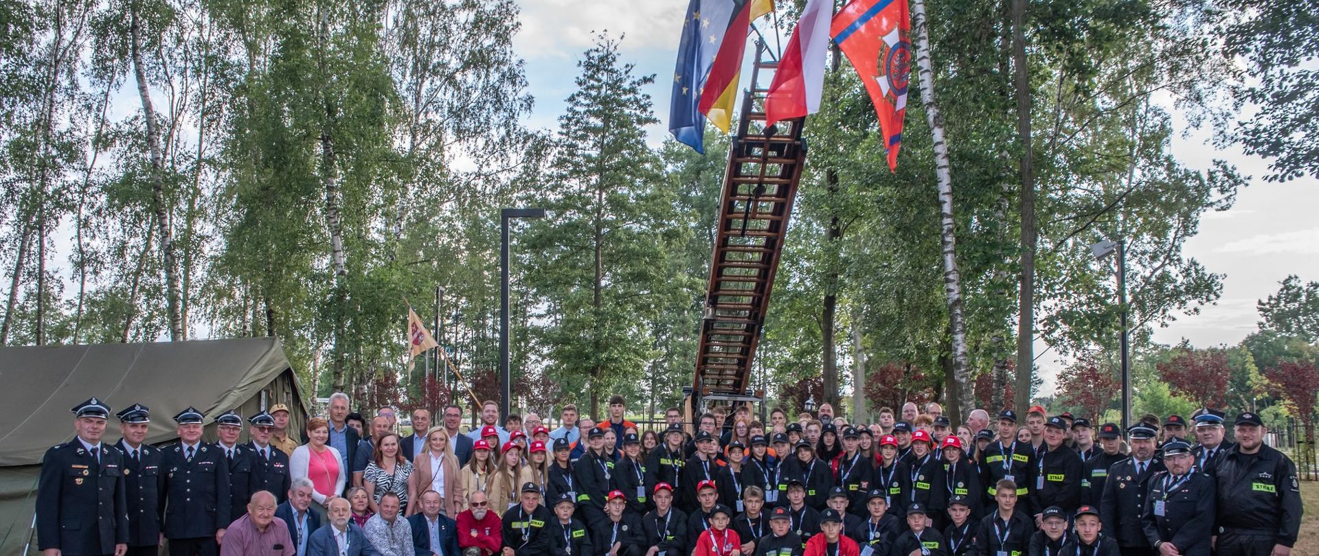 Zdjęcie przedstawia uczestników obozu Młodzieżowego oraz zaproszonych gości podczas oficjalnego otwarcia. Młodzież na zdjęciu w uniformach strażackich dedykowanych dla MDP.