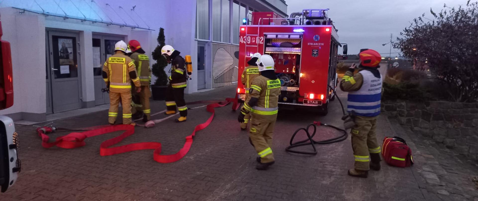 Strażacy przygotowują się do wejścia do budynku. W tle stoi pojazd pożarniczy.