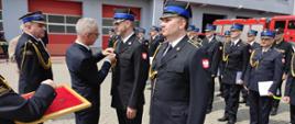 Zdjęcie przedstawia strażaków, którzy stoją na placu przed komendą podczas uroczystości obchodów dnia strażaka. Zastępca Prezydenta Miasta Konina wręcza strażakowi medal zasłużony dla miasta Konina. 