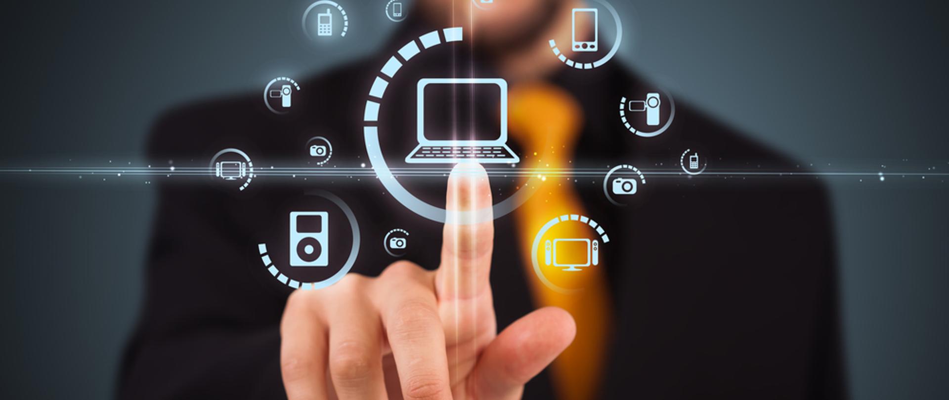 Piktogramy przedstawiające komputer, kamerę, laptop, telefon, osoba za piktogramami wskazuje piktogram przedstawiający komputer.