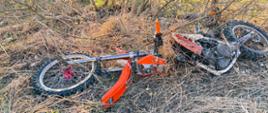 Na zdjęciu widoczny jest uszkodzony motocykl. Motocykl typu cross koloru pomarańczowego. Pojazd jest przewrócony, usytuowany na poboczu drogi.