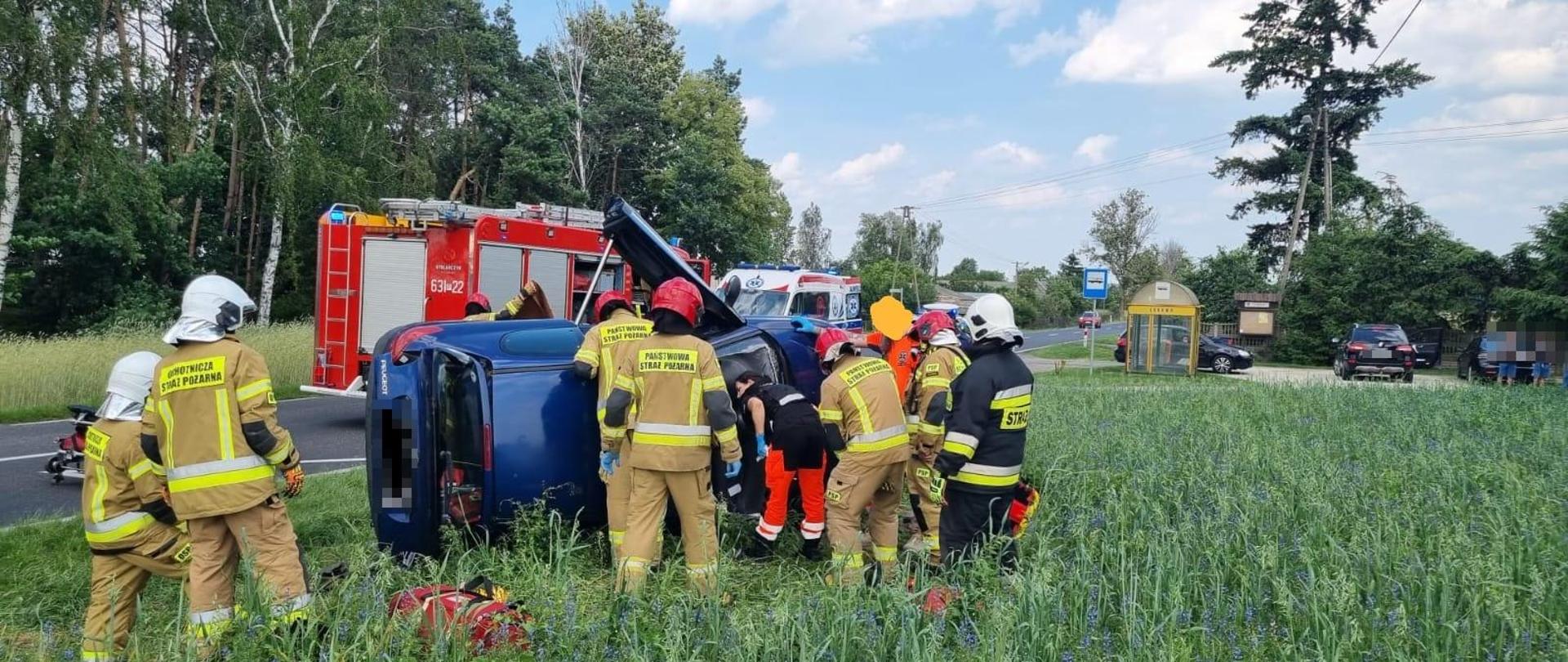 Na zdjęciu widać przewrócony na bok pojazd osobowy, pracujących strażaków oraz w tle wozy służb ratowniczych 