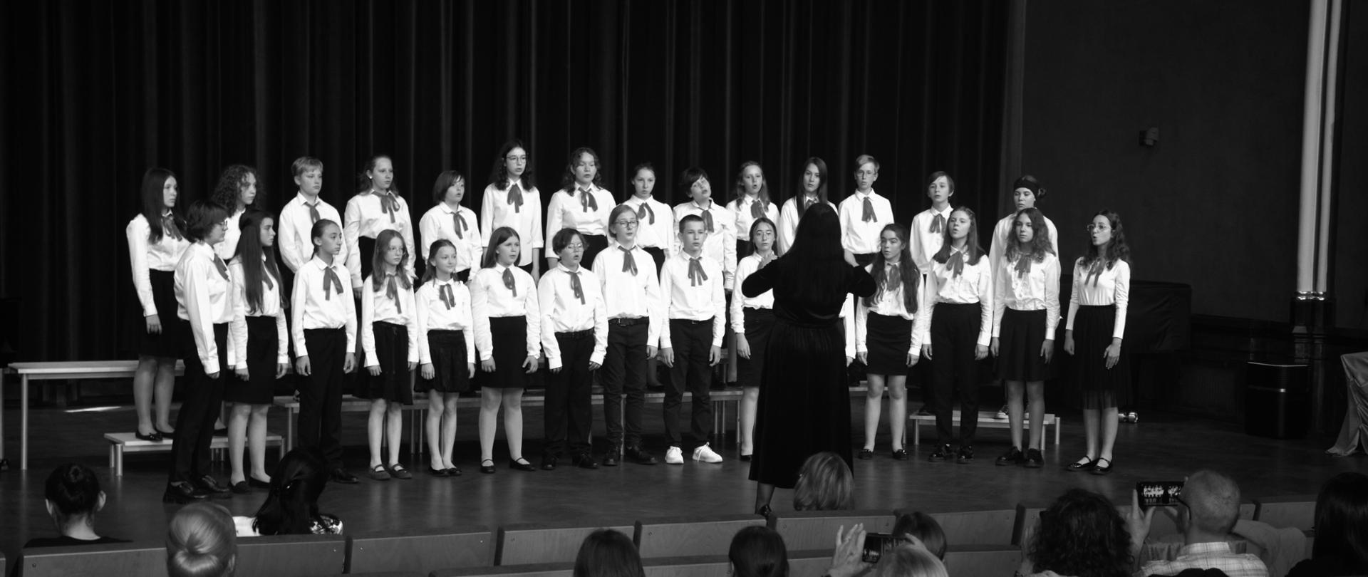 Zdjęcie z koncertu, młodzież (chór) ubrana na biało czarno stojąca frontem do słuchaczy, przed nimi pani dyrygent z podniesionymi rękami stojąca tyłem do słuchaczy